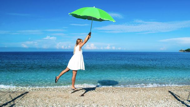 夏の晴れた青空と海を背景に小石のビーチの上に風によって彼女の手に緑の傘と白いドレスの若い女の子が提起されています. - 写真・画像