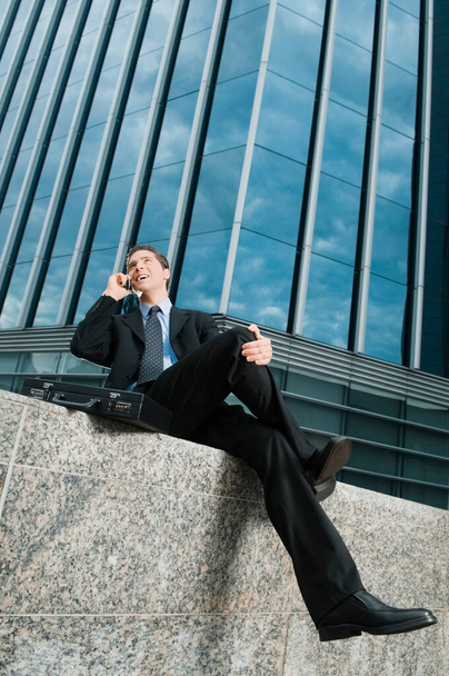 Low angle view of a businessman sitting at the edge of a wall and talking on a mobile phone - Foto, imagen