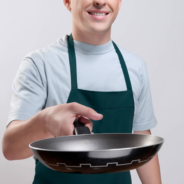 young man in apron holding a pot with a spoon on a white background - Φωτογραφία, εικόνα