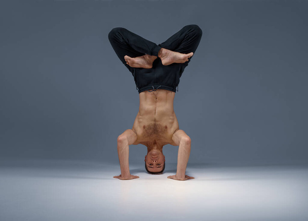 Male yoga stands on his head, hands and legs, meditation, grey background. Strong man doing yogi exercise, asana training, top concentration, healthy lifestyle - Photo, image