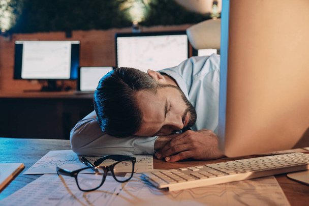 Exhausted young man sleeping at his working place while staying late in the office - Фото, изображение