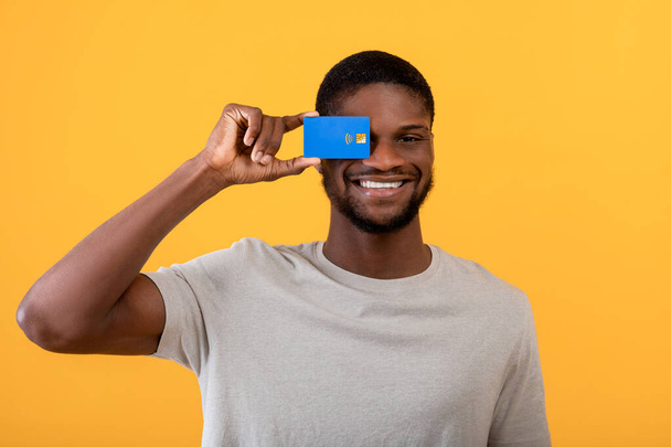 Emocionado tipo negro sosteniendo tarjeta de crédito en frente de su ojo, la promoción de la tarjeta bancaria en el fondo del estudio amarillo - Foto, Imagen