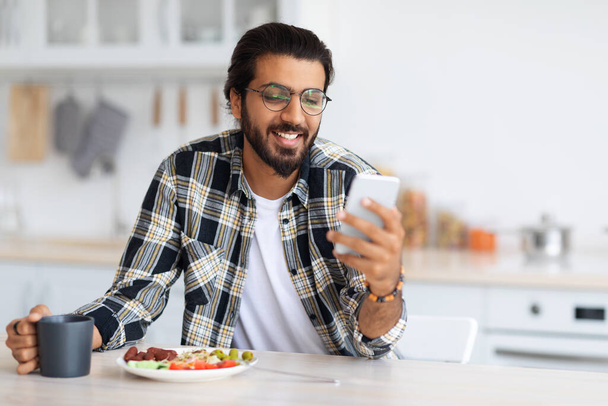 Uśmiechnięty mężczyzna z Bliskiego Wschodu jedzący zdrowe jedzenie i korzystający z telefonu - Zdjęcie, obraz