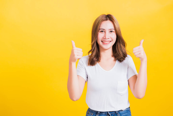 Portrait Asian Thai beautiful happy young woman smiling wear white t-shirt standing successful woman giving two thumbs up gesture sign in studio shot, isolated on yellow background with copy space - Foto, imagen