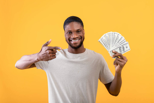 Big financial luck and profit concept. African american man holding money, pointing finger at it, yellow background - Photo, image