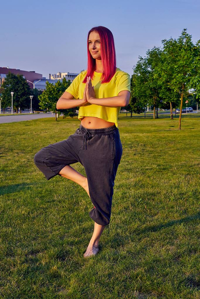young woman with pink hair trains yoga in a city park in a tree pose - Photo, Image