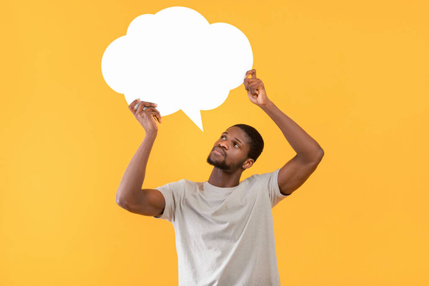 Thoughtful black man holding empty speech bubble over yellow studio background, free space for your ad design - Foto, Imagem