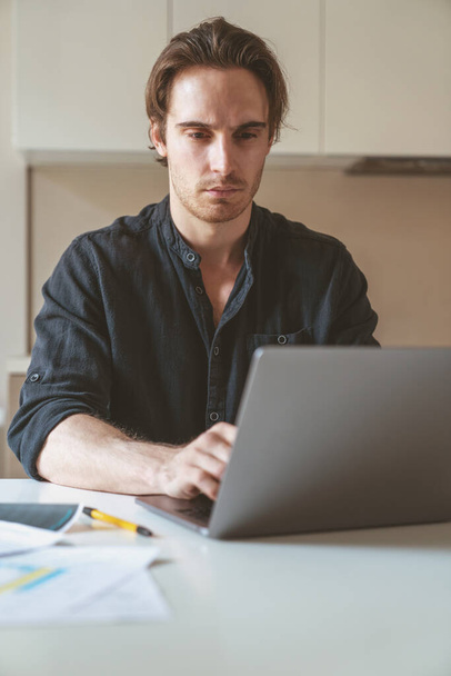 若い成功したビジネスマンは、ノートパソコンとコーヒーを飲んでテーブルのキッチンに座っています - 写真・画像