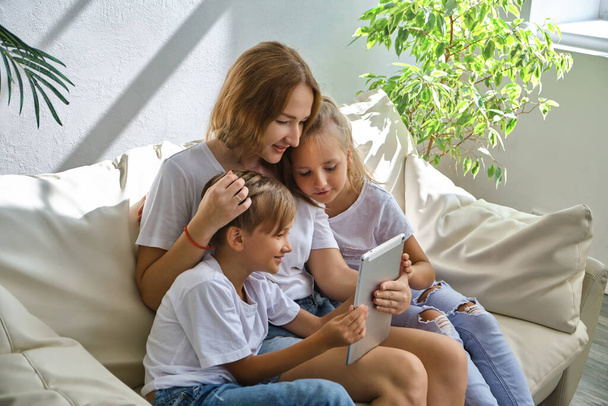 Fiatal anya a kisfiával és lányával Beszéljen videokommunikáción keresztül egy tablettel a kanapén otthon - Fotó, kép