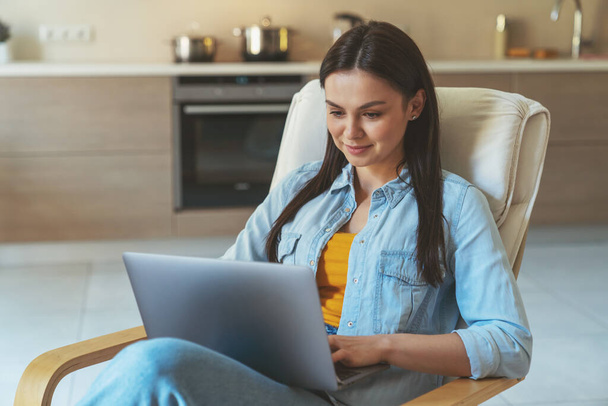 Jovem mulher empresária freelancer sentado em casa na cozinha e trabalhando usando laptop. - Foto, Imagem