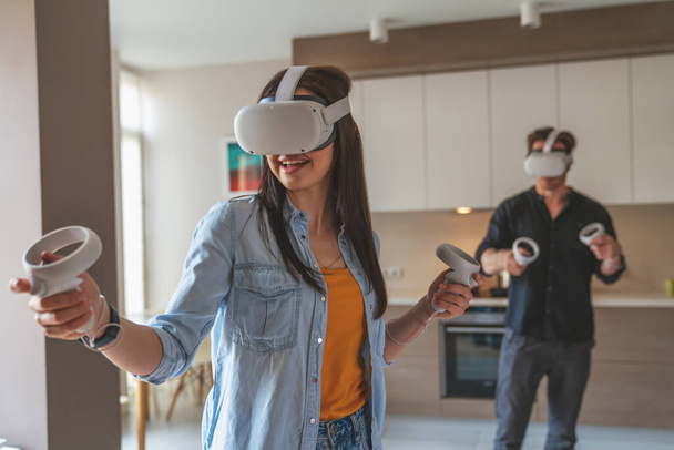 jong vrolijk paar man en meisje tijdens het spelen in virtual reality bril met controllers in handen thuis in appartement. - Foto, afbeelding