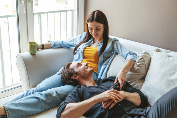 Young happy couple guy and girl hugging lying together on the couch at home in the apartment holding a remote control and having fun discussing something watching TV. - 写真・画像