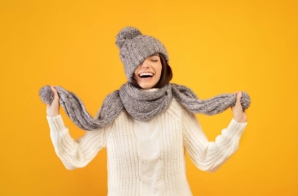 Overjoyed young lady in warm winter clothes pulling down knitted hat and playing with scarf on yellow background - Foto, Bild