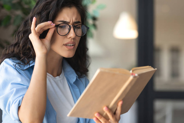 Beautiful young woman in glasses reading book, having poor eyesight at home - Foto, Imagem