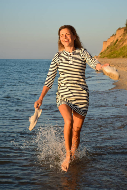 Deniz kıyısında, mavi gökyüzüne karşı, sarışın bir kız suyun üzerinde koşuyor, gülümsüyor ve elinde ayakkabıları tutuyor. - Fotoğraf, Görsel