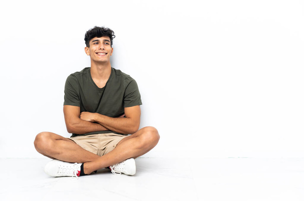 Mladý argentinský muž sedí na podlaze a pózuje s rukama v bok a usmívá se - Fotografie, Obrázek