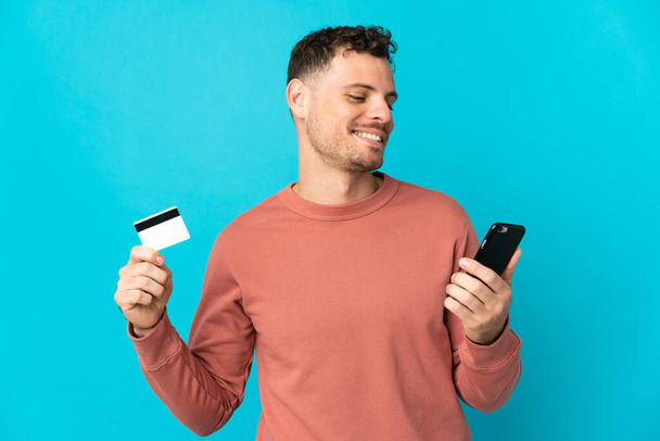 Young caucasian handsome man isolated on blue background buying with the mobile with a credit card - Zdjęcie, obraz