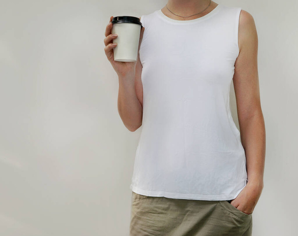 Close up of young girl in white tank top holding blank take away coffee cup in her hands over light gray background. Mockup of coffee to go cup and t-shirt print. - Valokuva, kuva