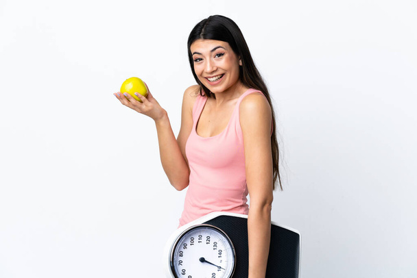 Young brunette girl over isolated white background with weighing machine and with an apple - Фото, изображение
