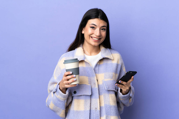 Joven morena sobre aislado fondo morado sosteniendo café para llevar y un móvil - Foto, Imagen