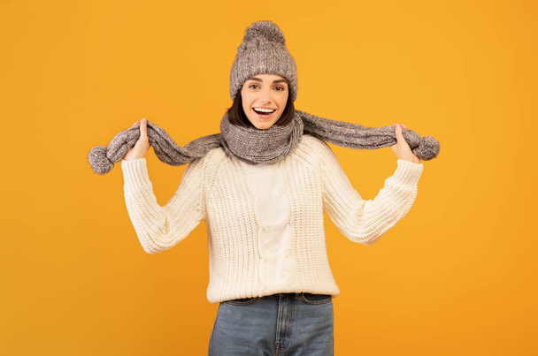 Winter fashion cocnept. Positive young woman wearing warm sweater, grey knitted hat and scarf, orange background - 写真・画像
