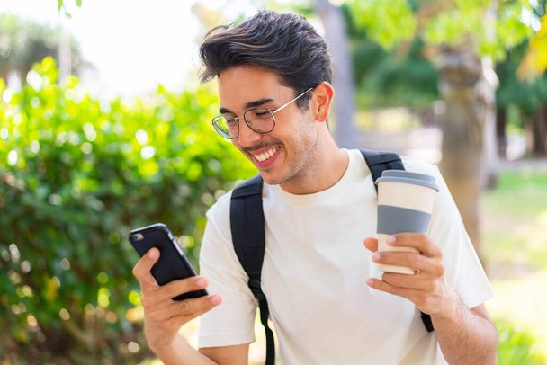 Jeune étudiant homme à l'extérieur en utilisant un téléphone portable et tenant un café à emporter - Photo, image