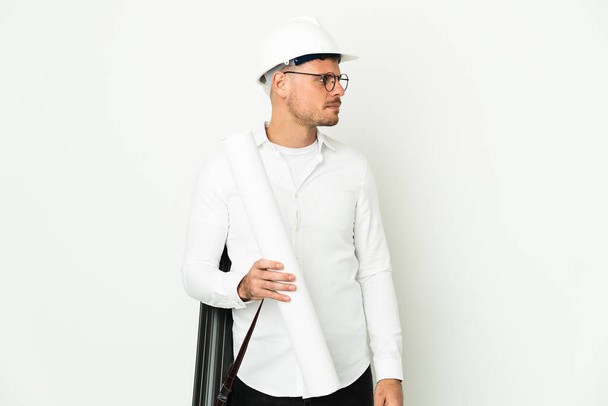 Giovane architetto uomo con casco e tenendo cianografie isolate su sfondo bianco guardando lateralmente - Foto, immagini