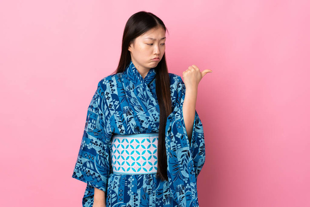Menina chinesa jovem vestindo quimono sobre fundo isolado infeliz e apontando para o lado - Foto, Imagem