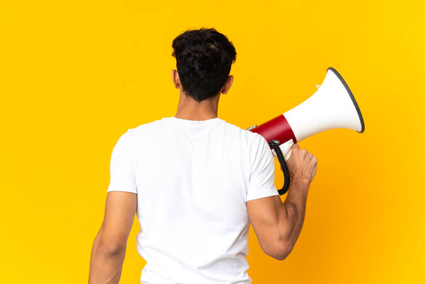 Young Argentinian man isolated on yellow background holding a megaphone and in back position - Photo, Image