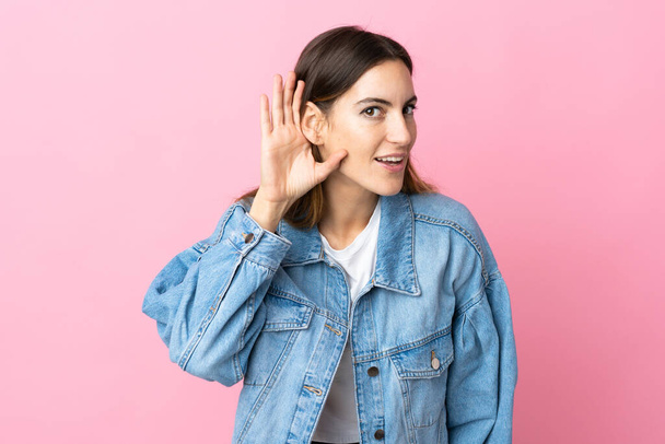 Young caucasian woman isolated on pink background listening to something by putting hand on the ear - Foto, immagini