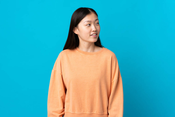 Menina chinesa jovem sobre isolado fundo azul olhando para o lado e sorrindo - Foto, Imagem