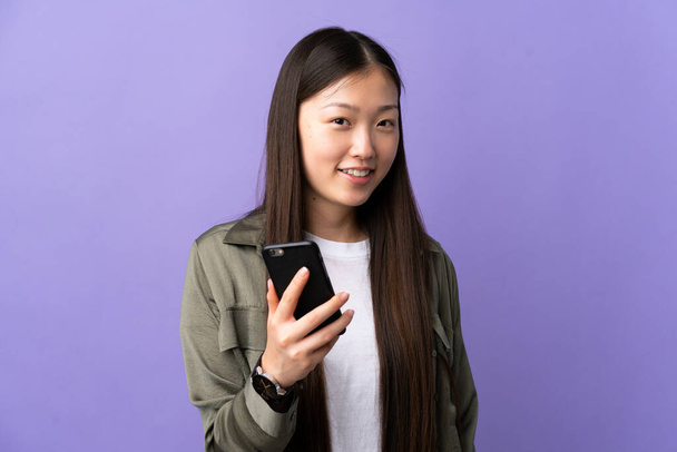 Молода китайська дівчина користується мобільним телефоном на ізольованому фіолетовому фоні з радісним виразом. - Фото, зображення