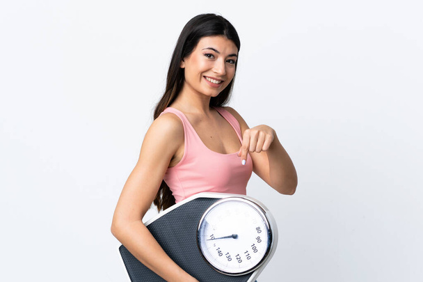 Young brunette girl over isolated white background holding a weighing machine and pointing it - Fotoğraf, Görsel