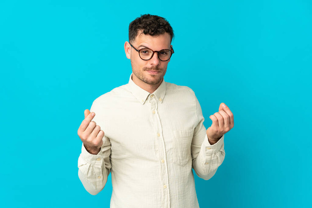 Young caucasian handsome man isolated on blue background making money gesture - Fotografie, Obrázek