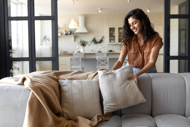 Lovely young woman putting soft pillows and plaid on comfy sofa, making her home cozy and warm, copy space - Foto, imagen