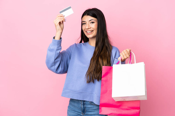 Νεαρή καστανή κοπέλα πάνω από απομονωμένο ροζ φόντο κρατώντας τσάντες για ψώνια και μια πιστωτική κάρτα - Φωτογραφία, εικόνα