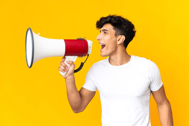 Young Argentinian man isolated on yellow background shouting through a megaphone to announce something in lateral position - Photo, Image