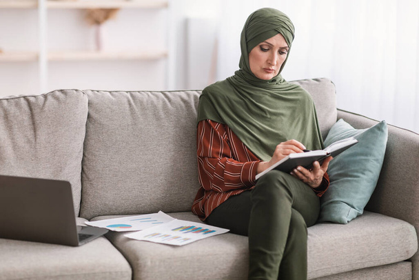 Зрелая мусульманская предпринимательница пишет бухгалтерский отчет, работая онлайн на дому - Фото, изображение