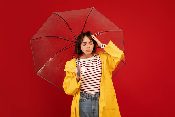 Millennial lady holding transparent umbrella and touching head, suffering from headache over red studio wall - Foto, imagen