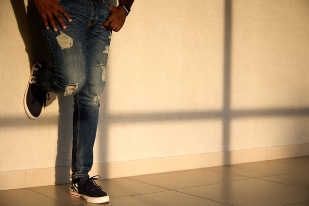 Прохолодний чорний чоловік в джинсах і кросівках стоїть на сонячному світлі перед вікном
 - Фото, зображення