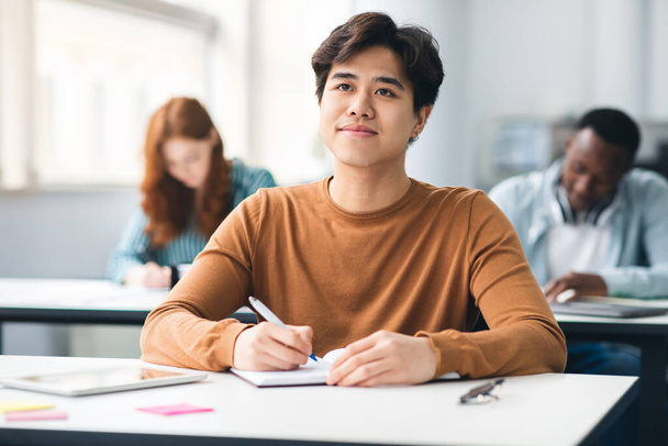 Χαμογελώντας ασιάτης φοιτητής κάθεται στο γραφείο στην τάξη - Φωτογραφία, εικόνα