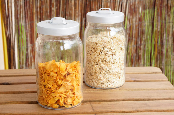 A closeup of two jars with oat-flakes and cornflakes on the wooden table - Фото, зображення