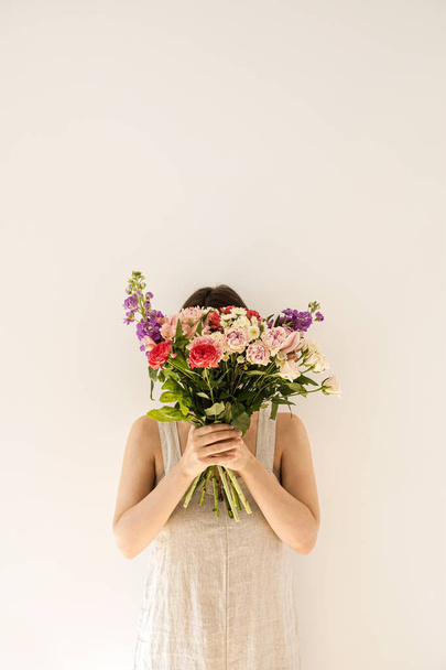 Young woman in beige washed linen dress hide her face by holding bouquet of colourful roses flowers against white wall. Beautiful wedding, Valentine's day holiday celebration floral composition - Φωτογραφία, εικόνα