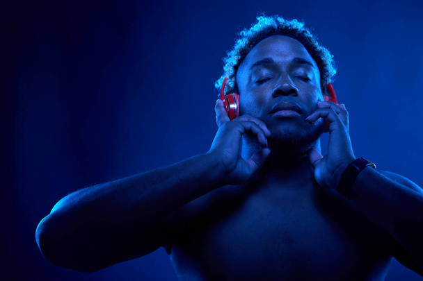 Uomo afro-americano in cuffie rosse senza fili ascolta musica e gode di musica su uno sfondo nero in raggi blu - Foto, immagini