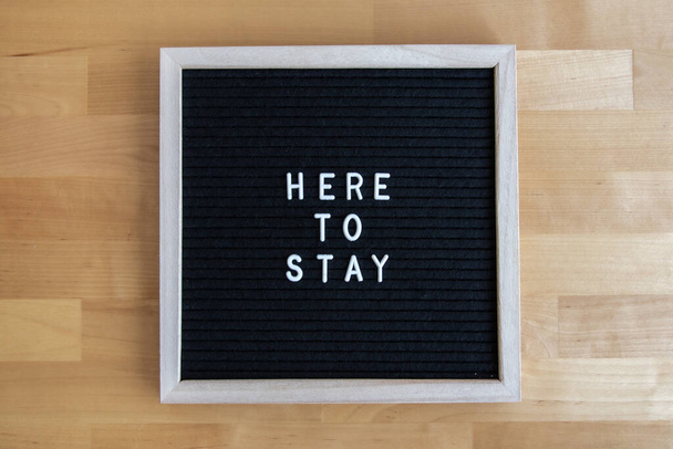 a top view shot of a blackboard with "here to stay" quote on it at a wooden table - Foto, Imagem