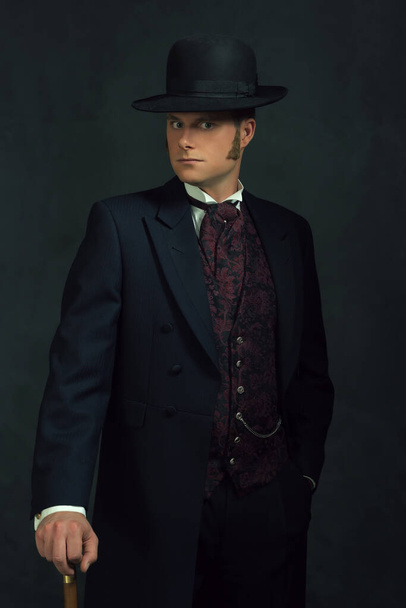 Mladý muž ve stylovém viktoriánském oblečení. Stojí s holí před tmavě šedou zdí. - Fotografie, Obrázek