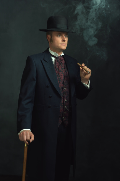 Cigar smoking young man in stylish vintage Victorian clothes with a walking stick stands in a room in front of a dark gray wall. - Foto, Bild