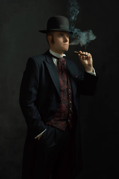 Πούρα που καπνίζουν νεαρός άνδρας σε κομψό vintage Βικτωριανά ρούχα στέκεται σε ένα δωμάτιο μπροστά από ένα σκούρο γκρι τοίχο. - Φωτογραφία, εικόνα