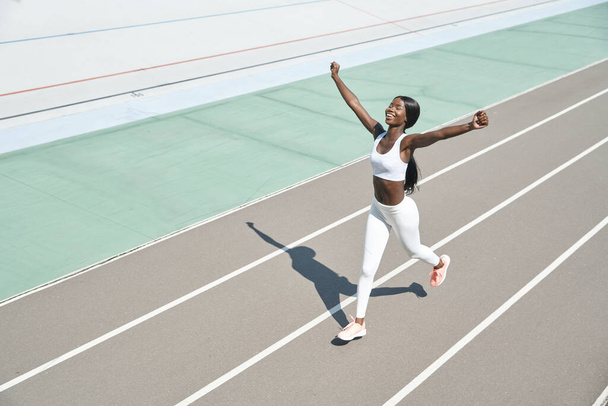 Top view of happy young African woman in sports clothing running on track and keeping arms raised - Photo, Image