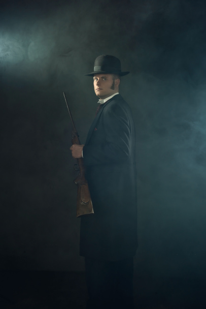 Shadowy portrait of a young man in vintage Victorian attire holding a rifle in a smoky room in front of a dark gray wall. - Fotografie, Obrázek